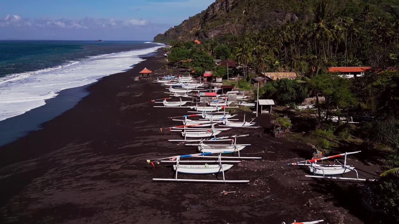 Traditional fishing boats rest on the black sand beach of Bali while rolling ocean waves meet the shore and green hills rise in the background captured in cinematic drone footage