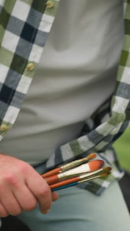 a close-up shot of an artist holding brushes, sitting on a chair in a grassy field, demonstrating a sitting pose. the checked shirt and outdoor setting emphasize the natural, creative atmosphere