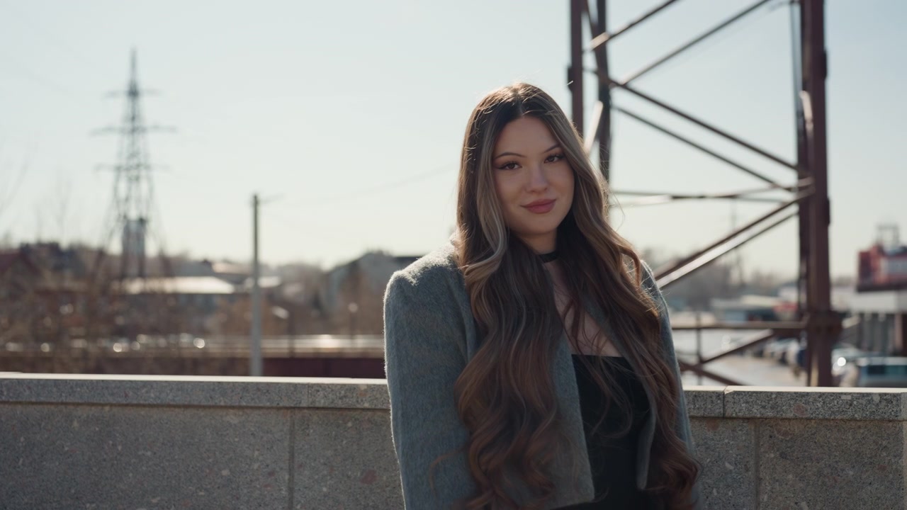 Bella influencer asiatica in stazione che indica lo skyline, con espressione sicura in un cappotto testurizzato, capelli lunghi illuminati dal sole, gesto coinvolgente e sorriso, con torri industriali e architettura urbana a fare da cornice.
