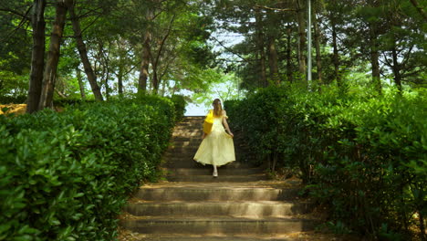 rear view of blonde woman in yellow summer dress with orange eco bag walking upstairs in green lush park in slow motion