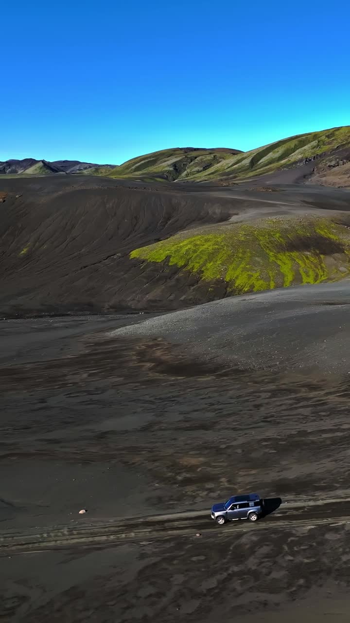 Ripresa verticale con drone di un fuoristrada che attraversa la piana vulcanica nera dell'Islanda, con colline muschiose e altipiani colorati, che cattura una scena di avventura overland remota.