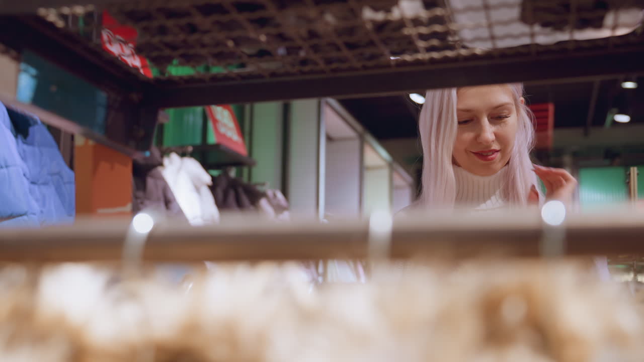 Rack view of young mother smiling while fixing eyes on clothing item she holds and inspects in well lit boutique setting with racks of jackets and coats in background evoking casual shopping mood