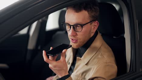 un joven apuesto en el auto está hablando mensajes de voz a través de un teléfono inteligente