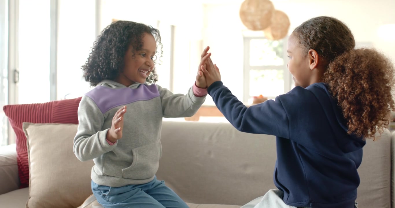 African American girls kneeling on couch raising hands playing clap game high-fiving near window