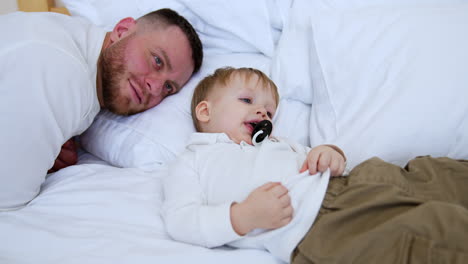 father and toddler son lie on the bed. calm kid is sucking a pacifier and smiles adorably.