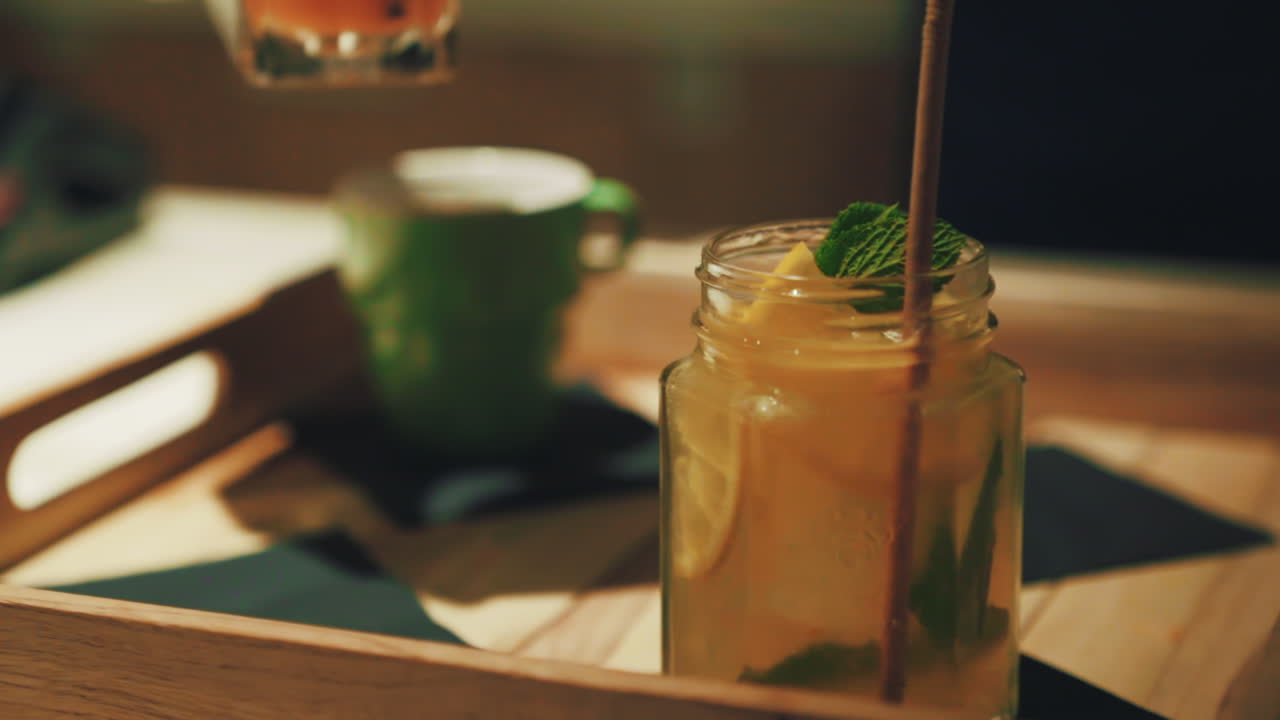 Hand putting a tall glass of fresh orange juice with a straw next to a jar of lemonade with mint and a green ceramic cup in the background on a wooden tray.
Push in close up.