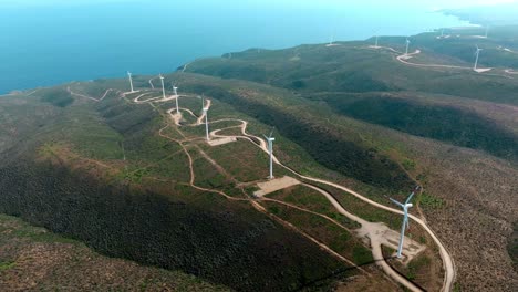 Vista-De-Seguimiento-Aéreo-De-Una-Línea-De-Molinos-De-Viento-En-Una-Montaña-Escarpada-Junto-Al-Océano