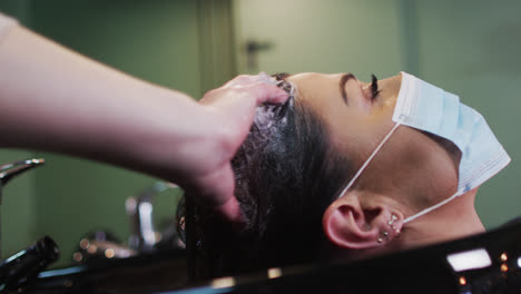 female hairdresser washing hair of female customer wearing face mask at hair salon