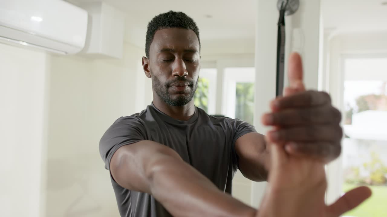 Mid adult African American male stretching wrist, clasping and pulling back in home gym with fan