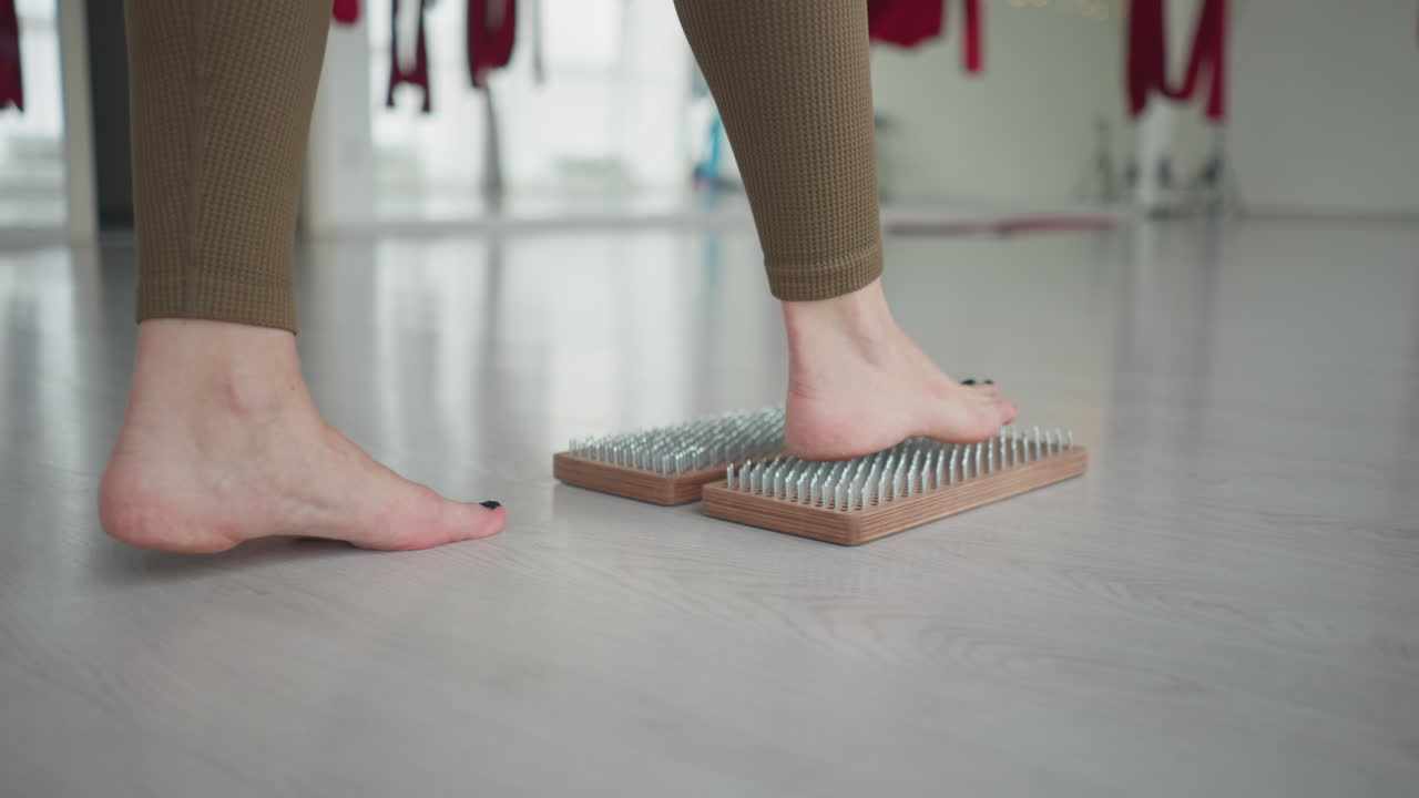Young woman barefoot practices acupressure stepping on wooden nail board in bright yoga studio with red aerial silks and large windows finding balance mindfulness and inner calm relaxation