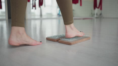 young woman barefoot practices acupressure stepping on wooden nail board in bright yoga studio with red aerial silks and large windows finding balance mindfulness and inner calm relaxation