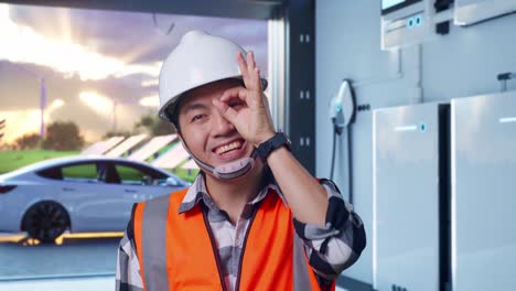close up of asian male engineer with safety helmet showing ok hand sign over eye and smiling to camera while standing with home energy storage system in a modern garage