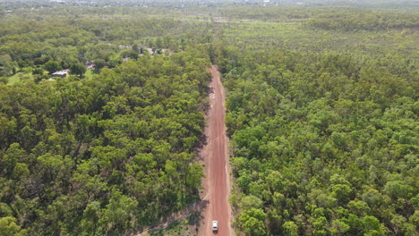 Toma-Aérea-De-Un-Dron-De-Movimiento-Lento-Y-Alto-De-Seguir-La-Conducción-De-Un-Automóvil-En-Una-Carretera-Roja-Larga-Y-Recta-Y-Matorrales-Verdes-Cerca-Del-Parque-Natural-De-La-Jungla-De-Holmes,-Darwin,-Territorio-Del-Norte