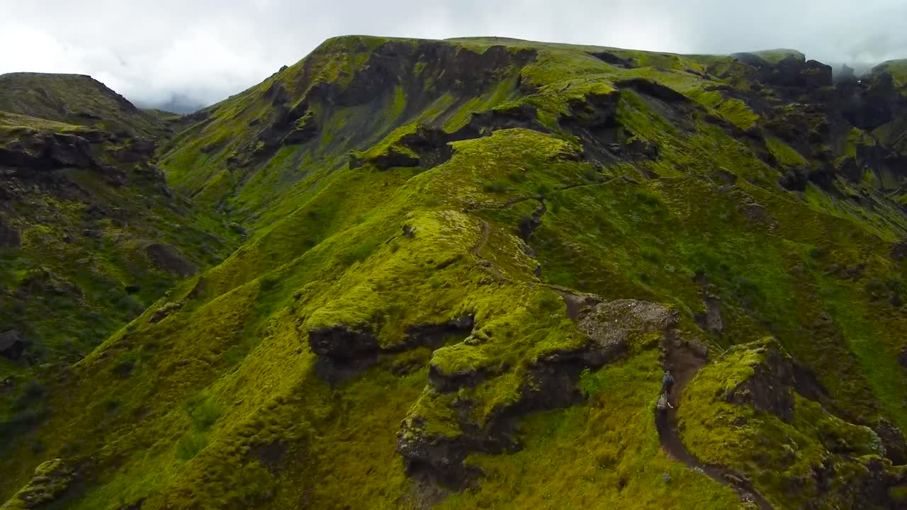 Aerial drone footage of Iceland green and steep mossy mountains during a cloudy day while two hikers are seen hiking and walking down on the path that goes up the mountains. Sharp edges and cliffs.