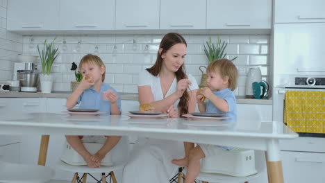 young family mom and two sons sitting in the kitchen at the white table eating burgers together and having fun. kids laugh at the dinner table