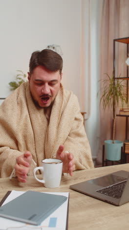 hombre enfermo freelance trabajando en una computadora portátil envuelto en cuadros sentado temblando por el frío bebiendo café caliente