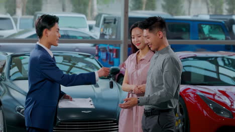 happy couple with attractive auto dealer in a suit presenting new car model to his client, shake hands with salesman and take key to new car