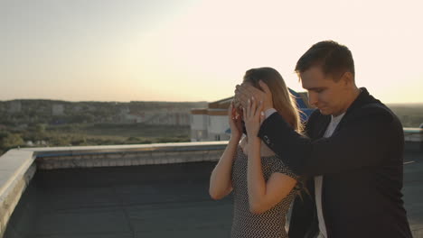 A-date-on-the-roof-a-man-is-leading-girl-closing-hands-eyes-preparing-romantic-surprise.-Show-a-beautiful-view-at-sunset.-The-woman-enjoys-the-surprise-and-smiles-looking-at-the-beautiful-view-and-the-man.