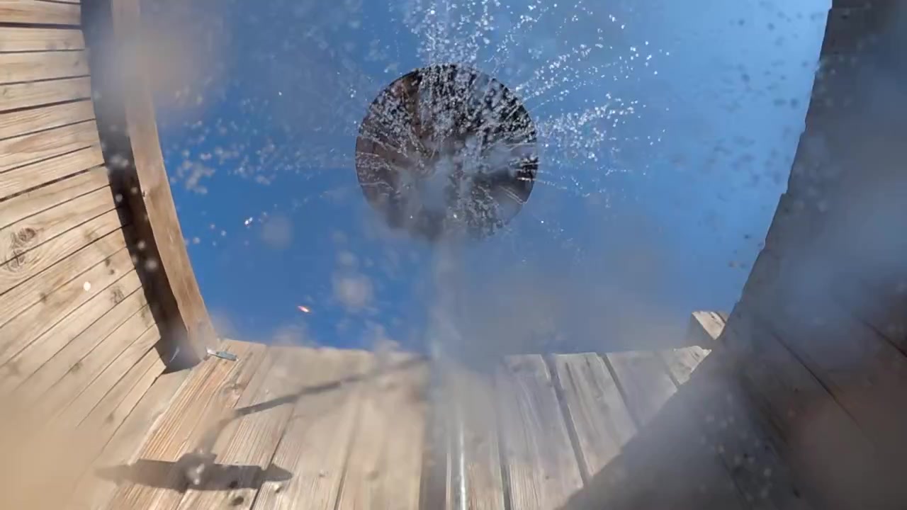 Ducha exterior desde un ángulo bajo, rociando agua refrescante del cabezal de la ducha contra un cielo azul claro y paredes de madera