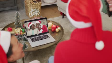 Diverse-family-with-santa-hats-using-laptop-for-christmas-video-call-with-happy-couple-on-screen