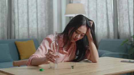 술에 취한, 우울한 아시아 여성이 테이블 위에 스마트폰을 들고 집에서 술을 마시기 전에 크 컵에 보드카를 부었습니다.