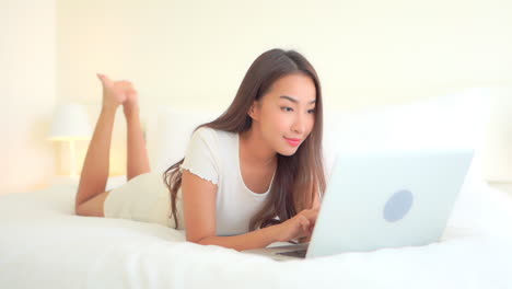 beautiful exotic woman typing on laptop keyboard, lying on bed in bright bedroom connecting with friends, full frame