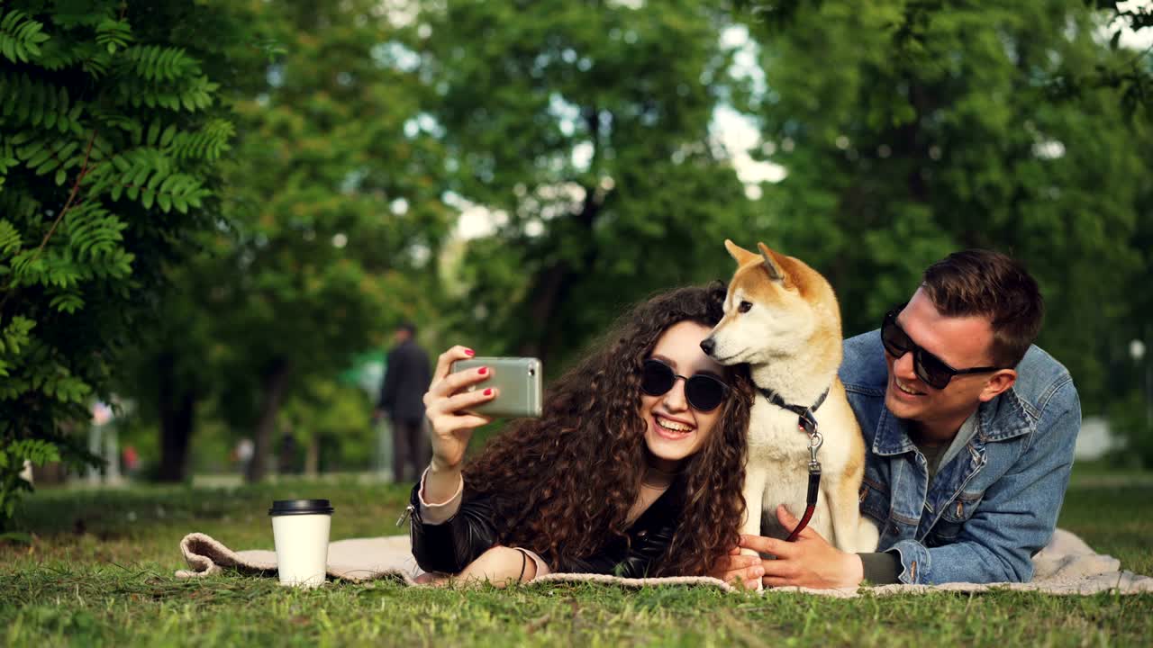 Young woman is taking selfie in the park lying on grass with her boyfriend and pet dog, adorable animal is sneezing and licking its nose, people are laughing.