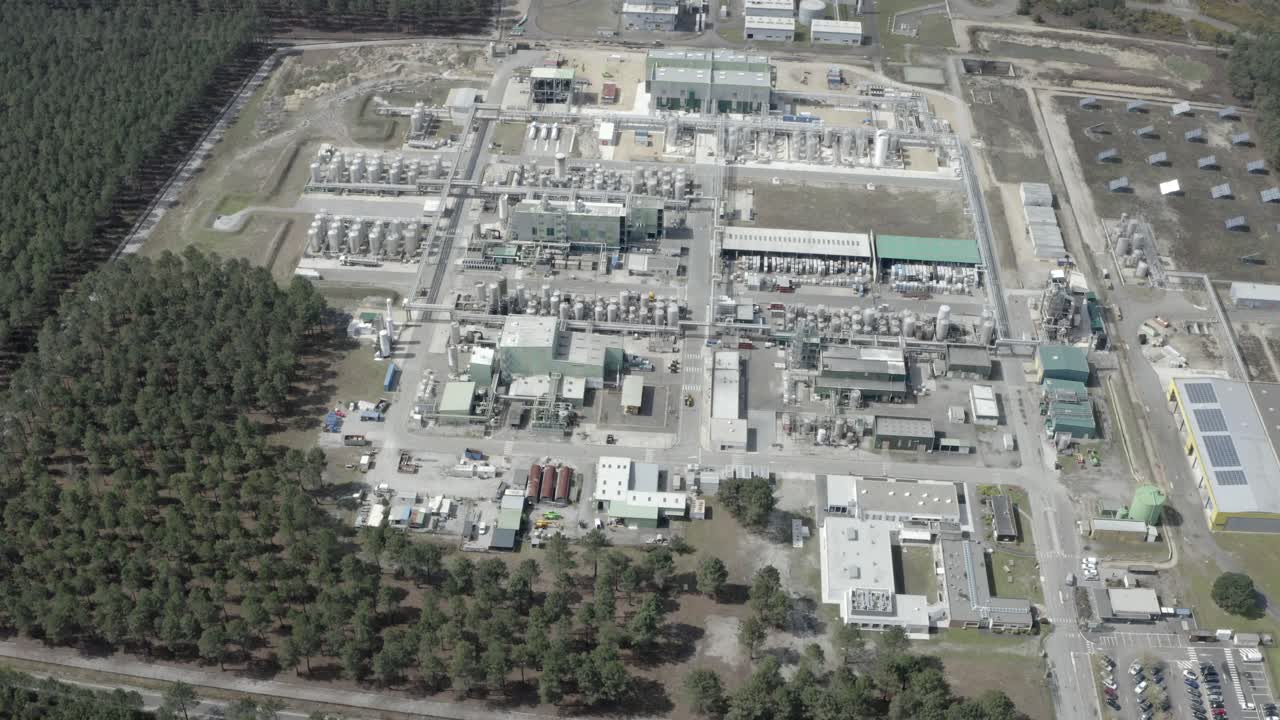 Aerial establishing shot of vast industrial complex nestled beside dense pine forest, showing buildings, tanks, and solar farm, Castets, France