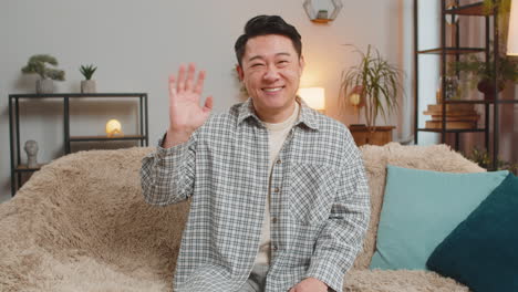 happy young man vlogger smiling friendly at camera and waving hands while sitting on sofa at home