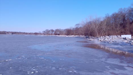 Backward-drone-flight-along-the-edge-of-a-frozen-river-with-broken-ice-along-the-edge-of-the-river-and-woods