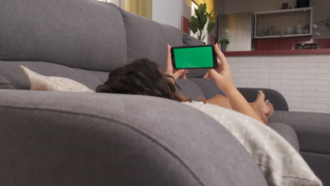 woman on the living room couch watching something on her cell phone, green screen on smart phone