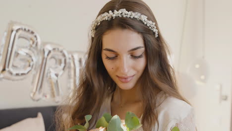 novia sosteniendo y oliendo ramo con banda para el cabello con flores, mirando a la cámara mientras sonríe