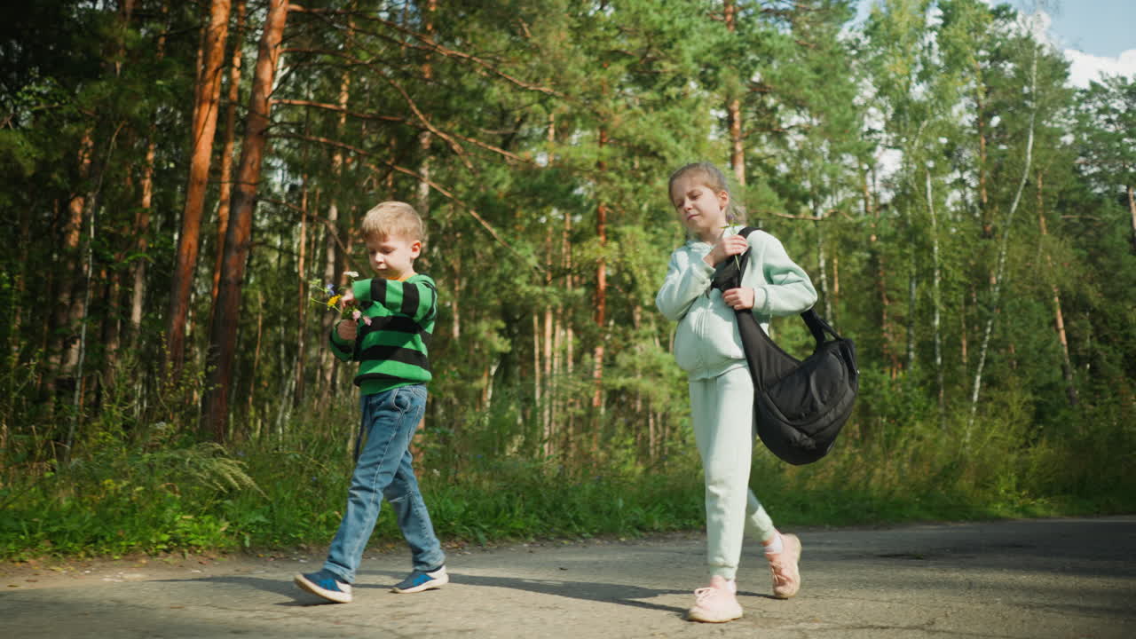 Children walking along forest road during daylight as girl adjusts shoulder bag and boy walks beside holding wildflower, surrounded by green trees, natural light, and serene outdoor environment