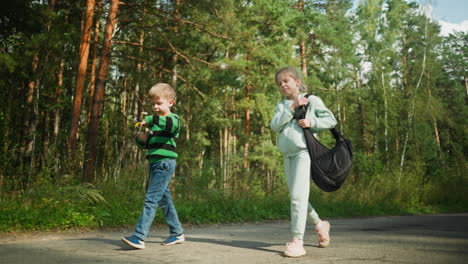children walking along forest road during daylight as girl adjusts shoulder bag and boy walks beside holding wildflower, surrounded by green trees, natural light, and serene outdoor environment