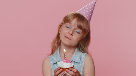 happy child girl kid celebrating birthday party, makes wish blowing burning candle on small cupcake