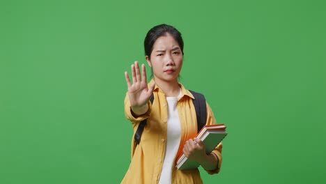 asian woman student with a backpack and some books shaking head and disapproving with no hand sign while standing in the green screen background studio