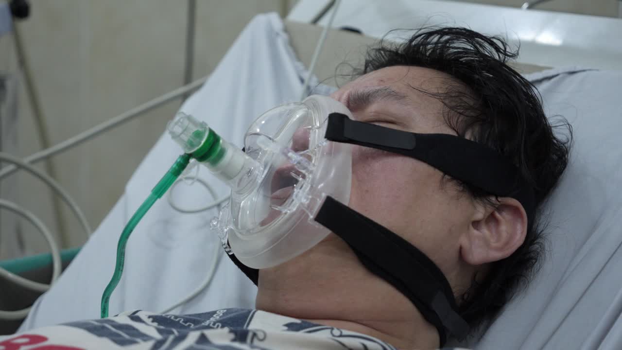 Paciente masculino com máscara de oxigênio em cama de hospital, atendimento de emergência médica.