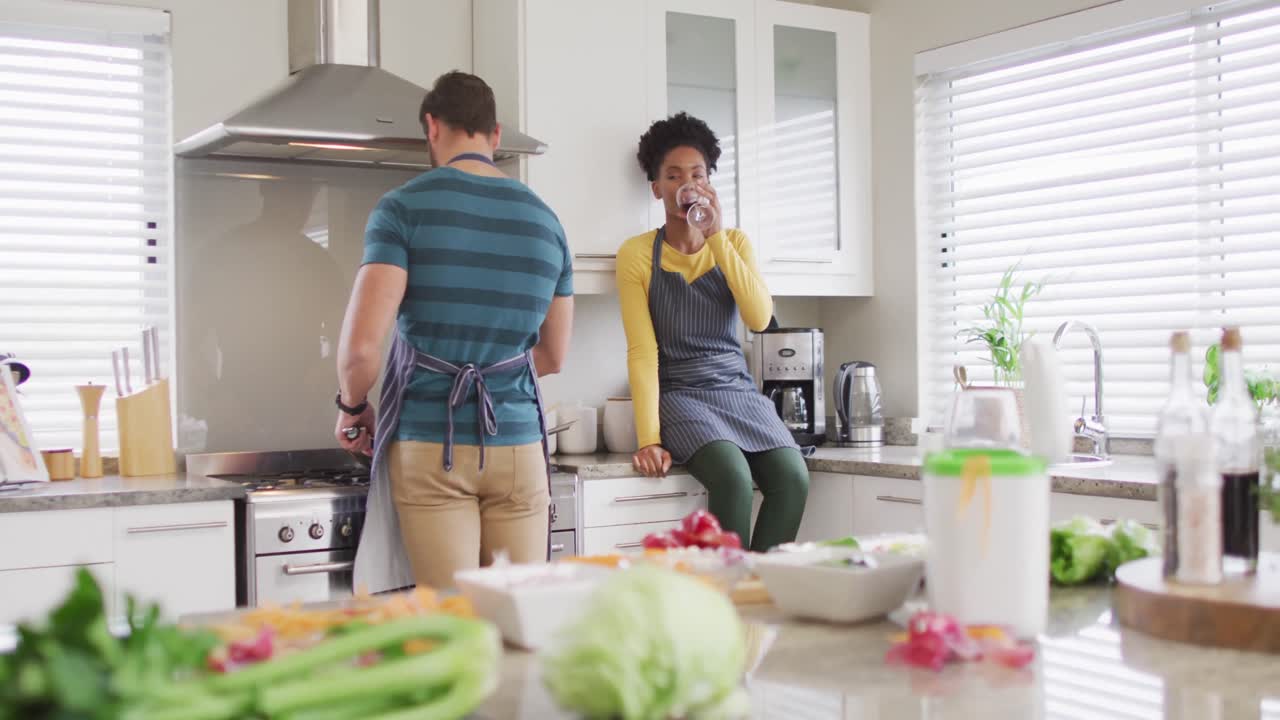 Vídeo de um casal diverso feliz preparando uma refeição, bebendo vinho e se divertindo na cozinha