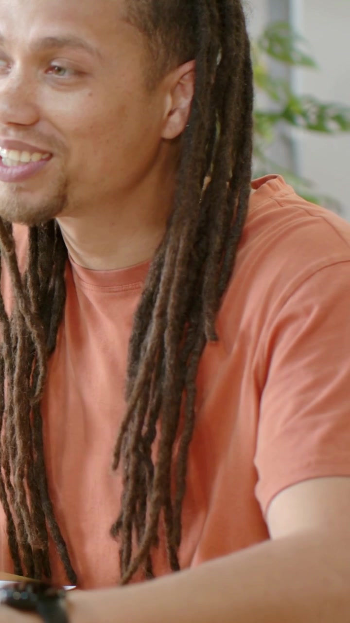 Video verticale: Inizio di una conversazione, uomo con i dreadlock in maglietta arancione che parla, dando un'occhiata all'orologio.