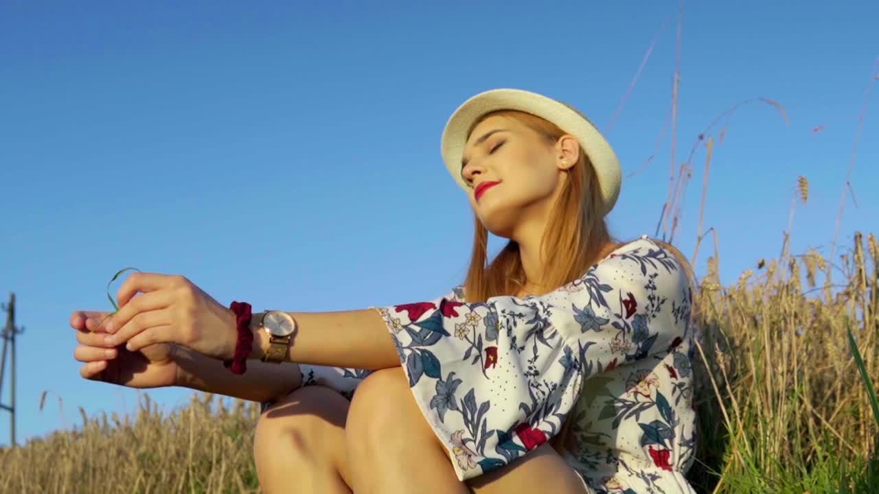 Stunning HD footage of a white caucasian girl in a dress with a knitted hat and red lipstick sitting in a wheat field, lost in thought, basking in the setting sun's warm embrace