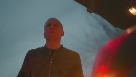 A man in a green jacket looks up with a tense expression while standing by an open car trunk at dusk, with a phone and gun in his hand as he close the car trunk