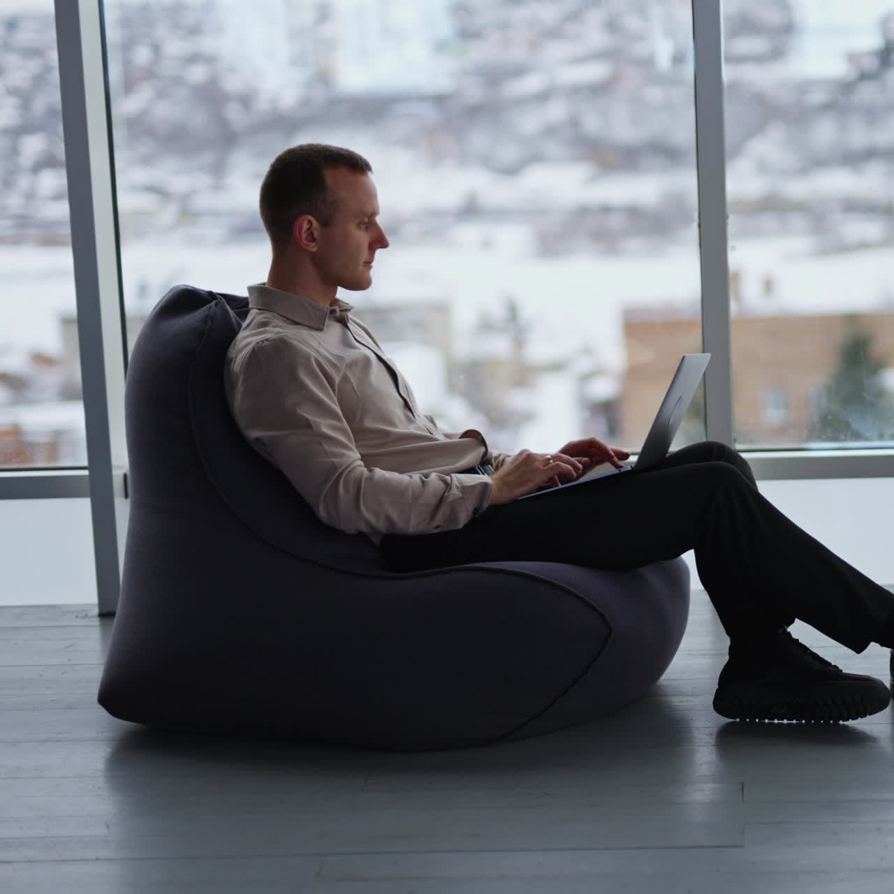 Freelance male businessman working at laptop in comfortable room. Man sitting in bean bag chair at backdrop of panoramic windows and winter cityscape in blur