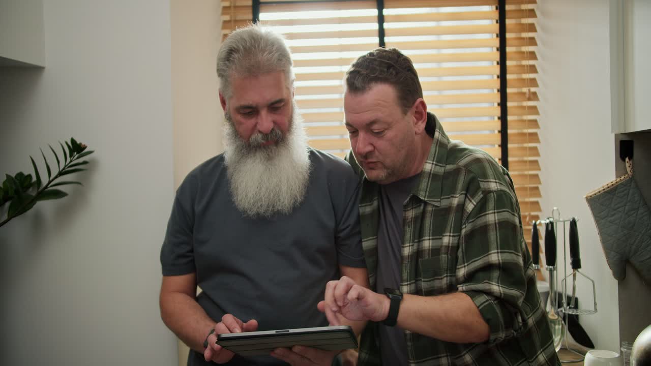 An interested middle-aged brunette man with stubble in a green checkered shirt together with his elderly boyfriend with a gray gray beard and a lush mustache are choosing something on their electronic tablet during the day at home