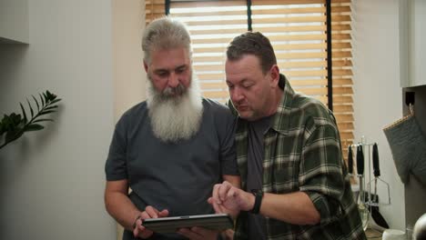un hombre moreno de mediana edad interesado con paja en una camisa a cuadros verdes junto con su novio mayor con una barba gris gris y un bigote exuberante están eligiendo algo en su tableta electrónica durante el día en casa