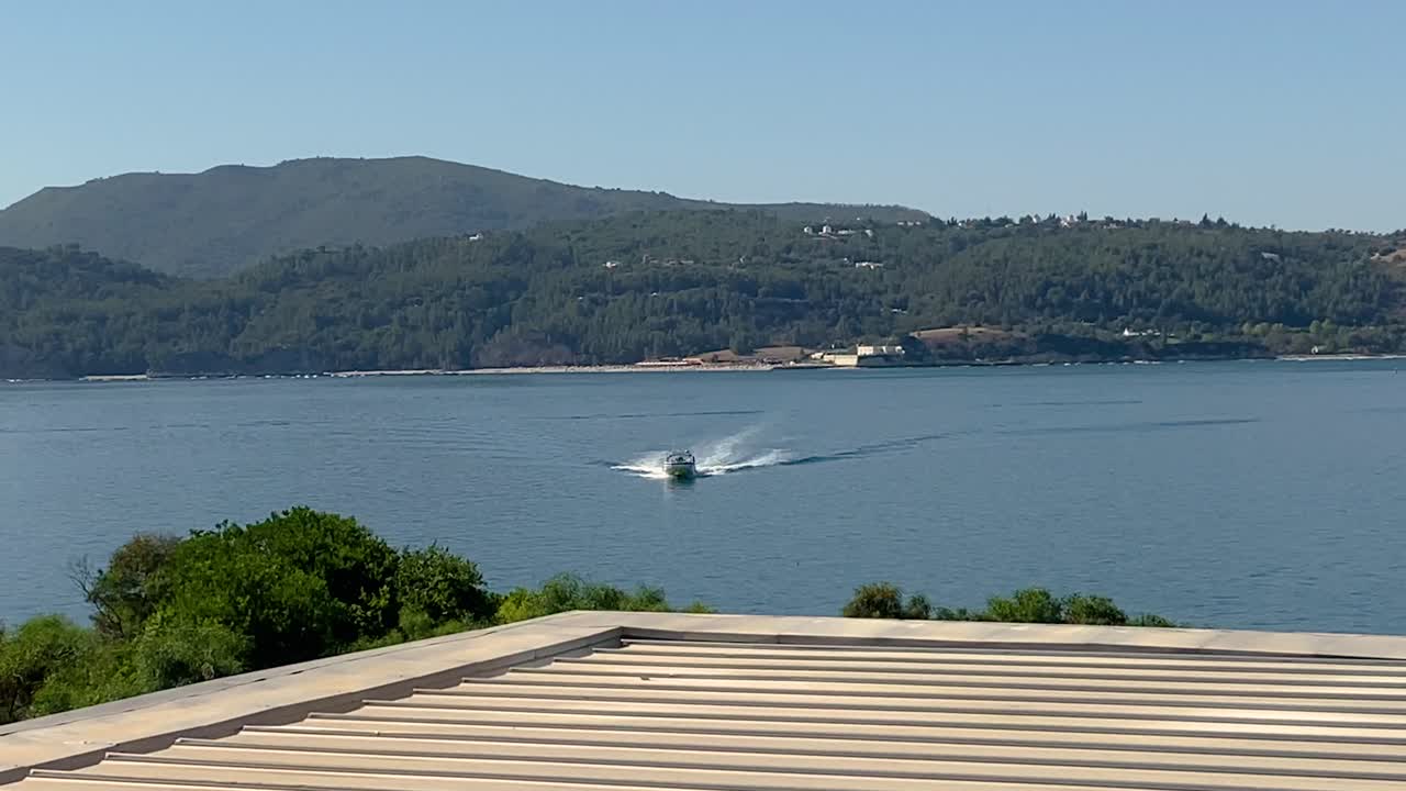 트로이아 섬의 푸른 녹색 바다를 따라 이동하는 선박 보트 요트 페리를 보여주는 텔레 줌 샷 배경에 녹색 산을 보여주는 나무가 있습니다.
