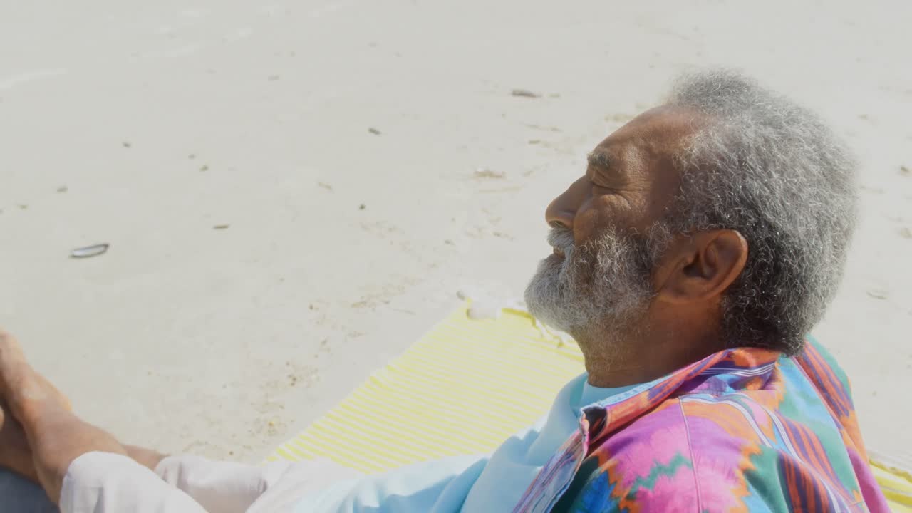 Vue latérale d'un homme afro-américain senior actif et pensif se relaxant sur la plage au soleil, 4K.