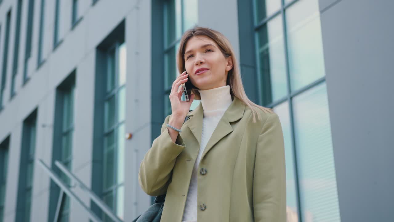 Smiling young woman stands in front of modern glass office building and having video call on mobile phone. Girl laughs and enjoys good news, communicating with friends outdoors