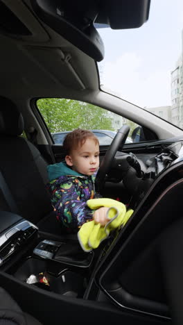 daddy's little helper sitting in the car. bab boy uses yellow rag to clean the panels in automobile. vertical video.