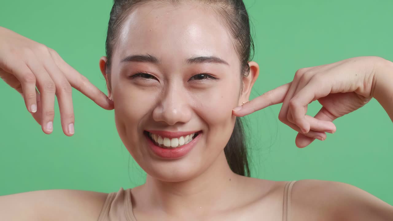 아름다운 아시아 여성이 인지손가락으로 얼굴을 만지고 스튜디오의 녹색 화면 배경에서 카메라를 보고 미소 짓고 있습니다.
