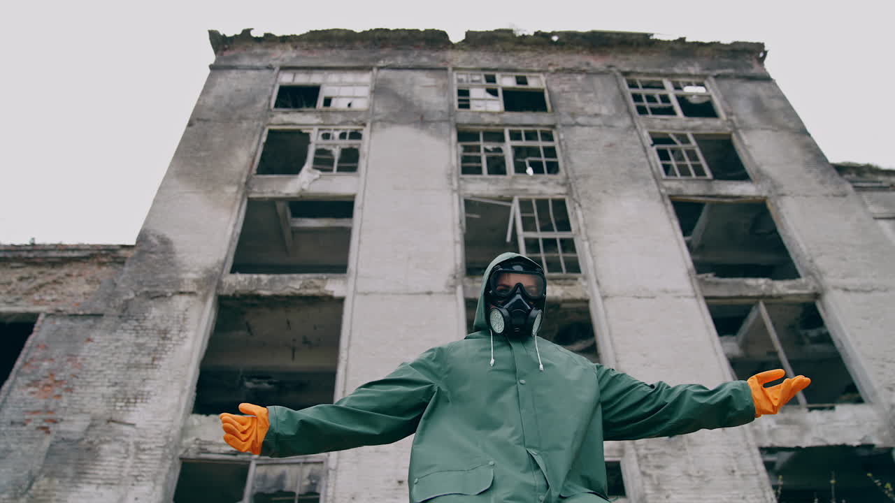 Abandoned place and a person in safety suit. Survivor in gas mask and hazmat suit standing with stretched arms on ruined building background. Chemical catastrophe.